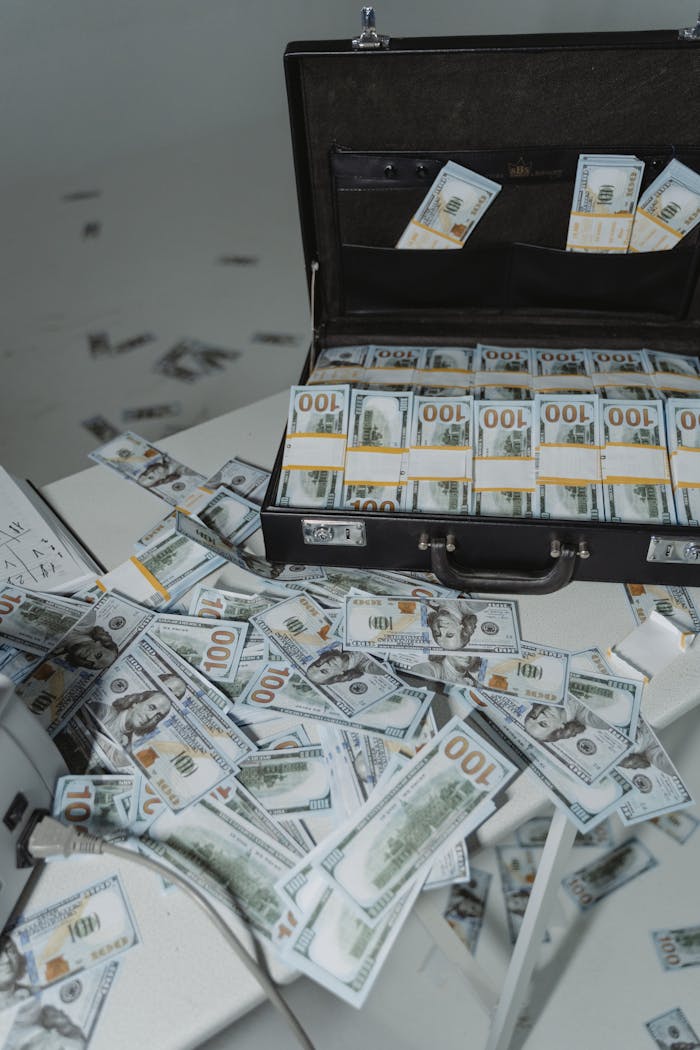A briefcase overflowing with neatly stacked US dollar bills surrounded by scattered currency on a table.
