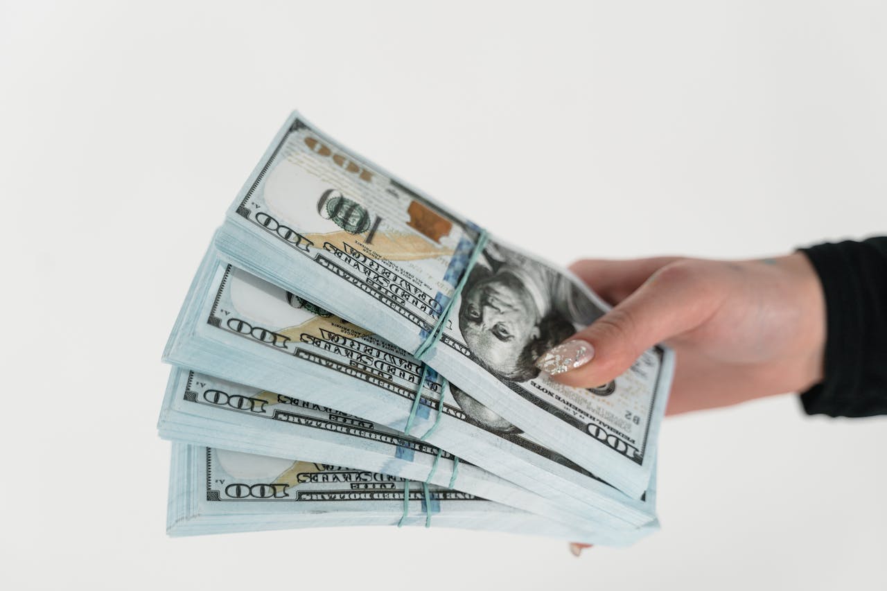 Close-up of a hand holding several stacks of US hundred dollar bills on a white background.